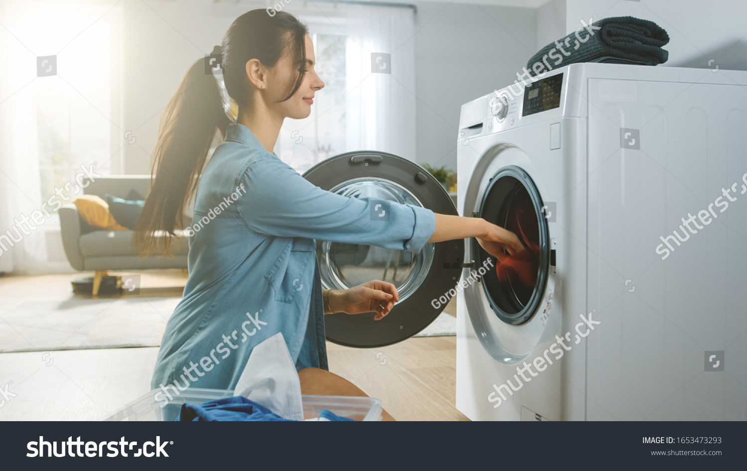 Beautiful Smiling Brunette Young Woman Sits in Front of a Washing Machine in Homely Jeans Clothes. She Loads the Washer with Dirty Laundry. Bright and Spacious Living Room with Modern Interior. #1653473293