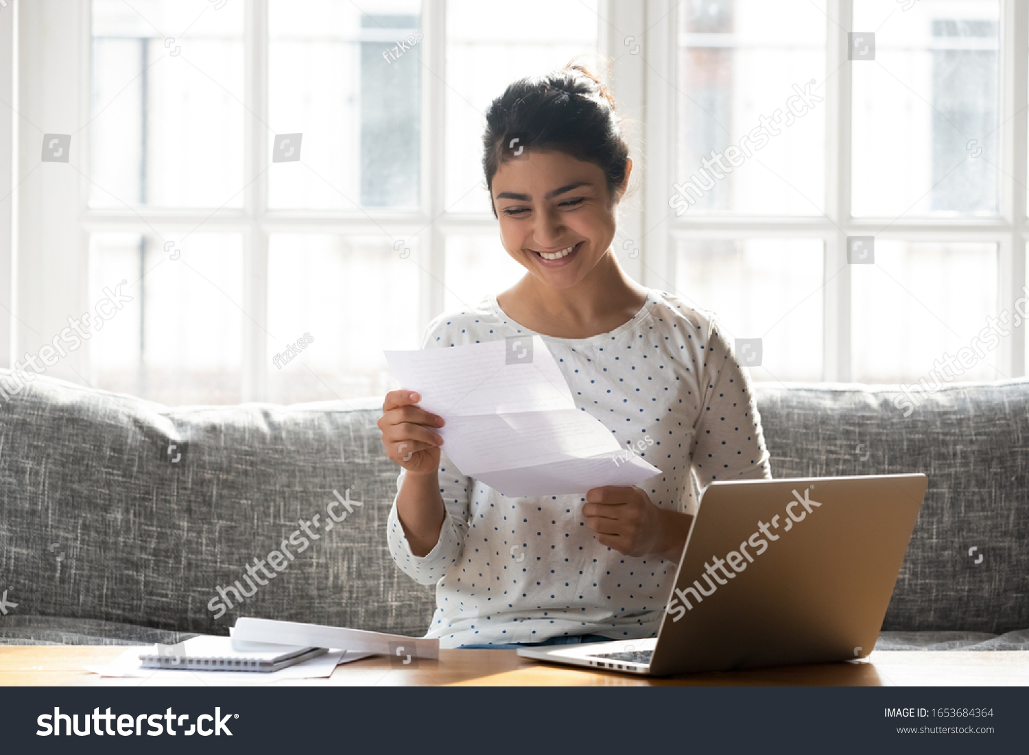 Indian ethnicity woman sitting on couch at home reading paper notice receive good news feels happy, cheerful student female looking at document enjoy exam results or college admission letter concept #1653684364