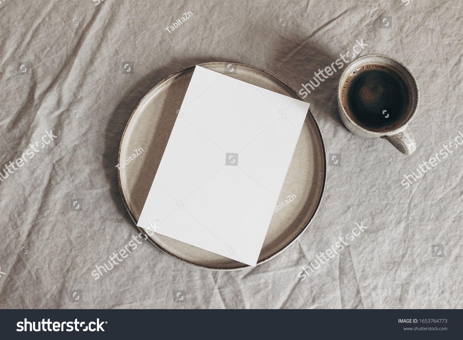 Cup of coffee and blank greeting card, invitation on ceramic plate. Moody breakfast table mockup scene. Beige linen tablecloth background. Sparse still life composition  Rustic flat lay, top view. #1653764773