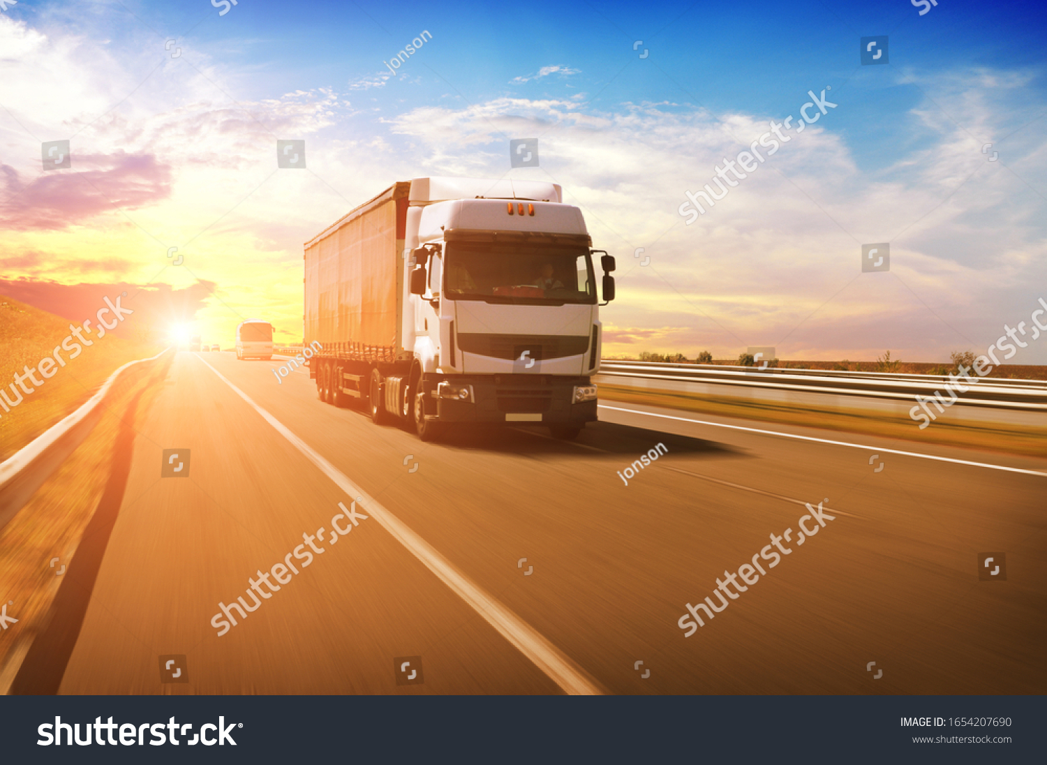 A big white truck with a red trailer and other cars on the countryside road in motion against a night sky with a sunset #1654207690
