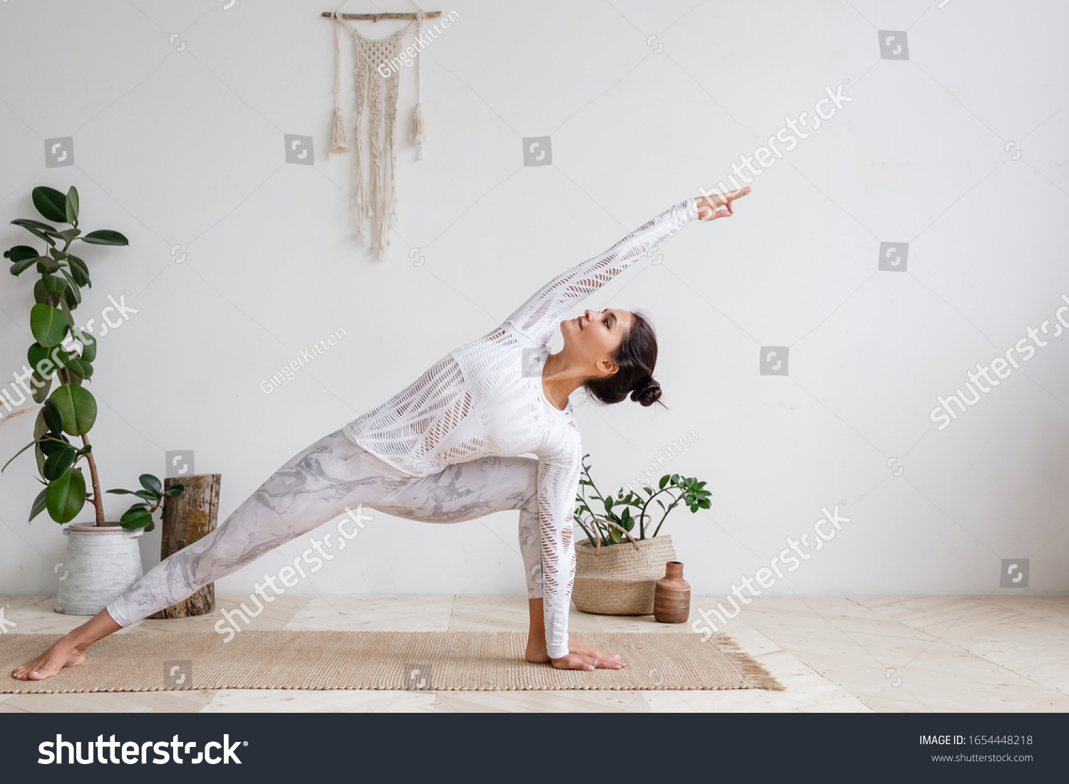 Side view of slim pretty positive young brunette woman doing Utthita parsvakonasana exercise, Extended Side Angle pose, on mat on floor surrounded by houseplants on white wall. Yoga and pilates #1654448218