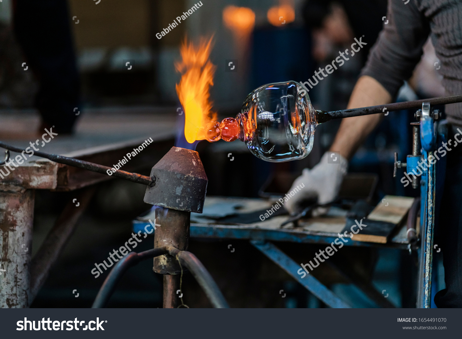Close-up of a glassblower artisan shaping the hot molten glass at strong fire inside his workshop. Manual glass processing by the craftsmen inside a glass factory in Transylvania, Romania.  #1654491070