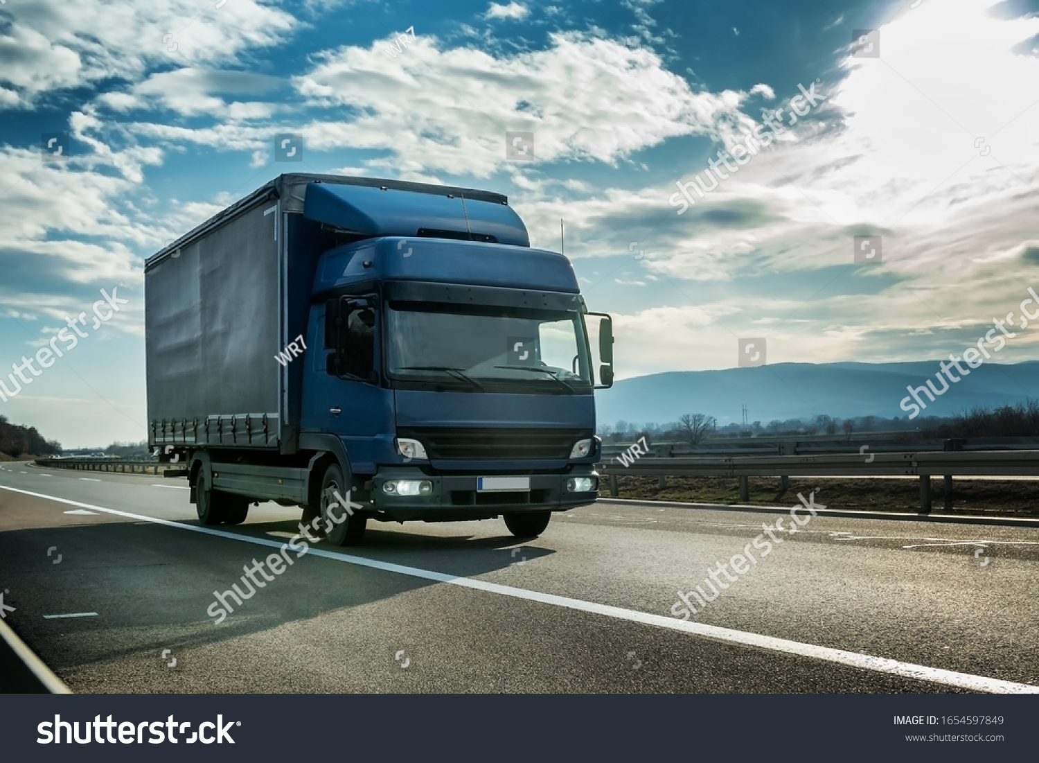 Blue semi trailer truck on a highway driving at bright sunny sunset. Transportation vehicle #1654597849