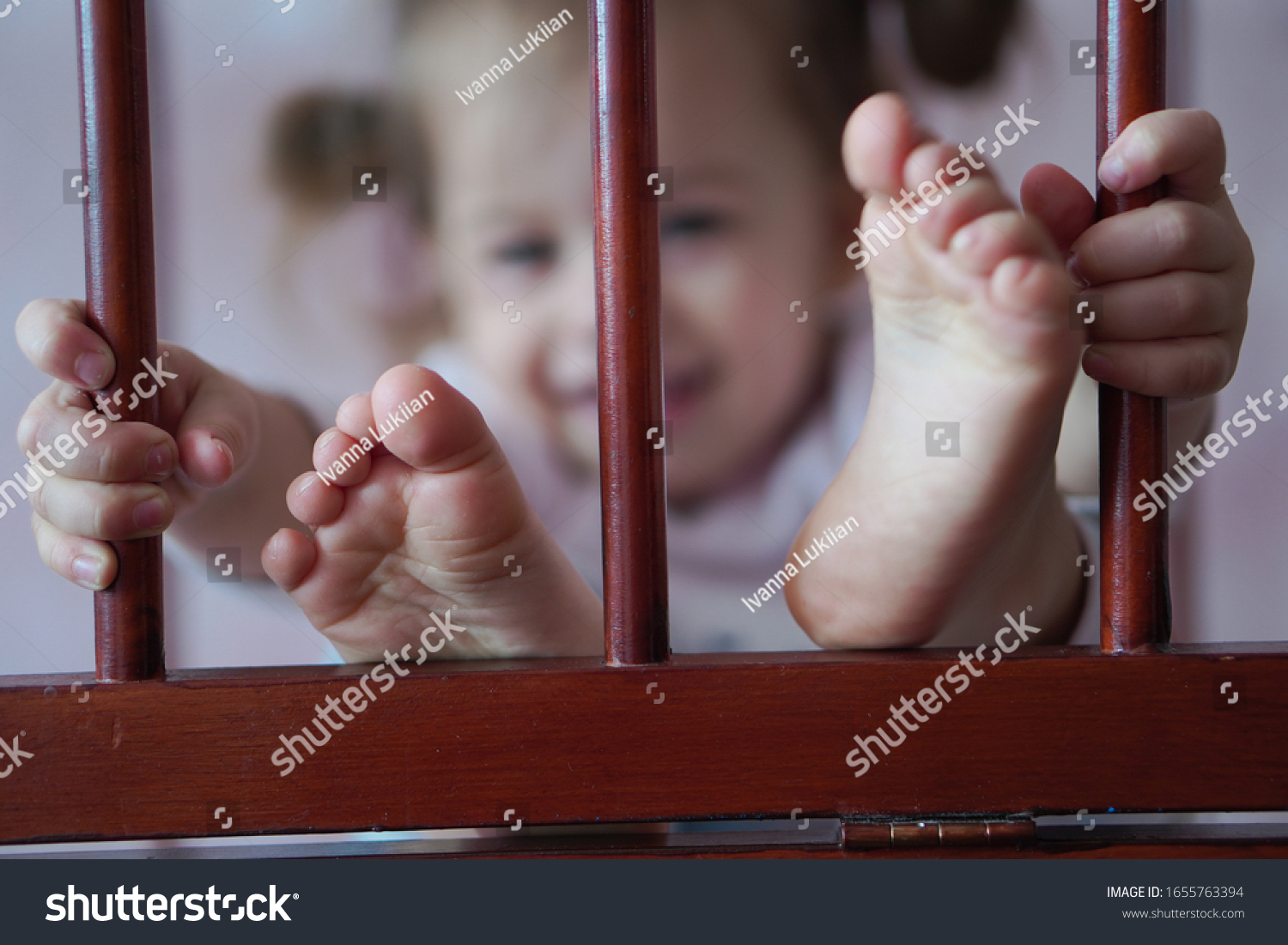 The little girl laughs and holds her hands behind the partitions in the cradle. Smiling girl looks from the cradle. The feet of a young child are pushed out of the cot #1655763394