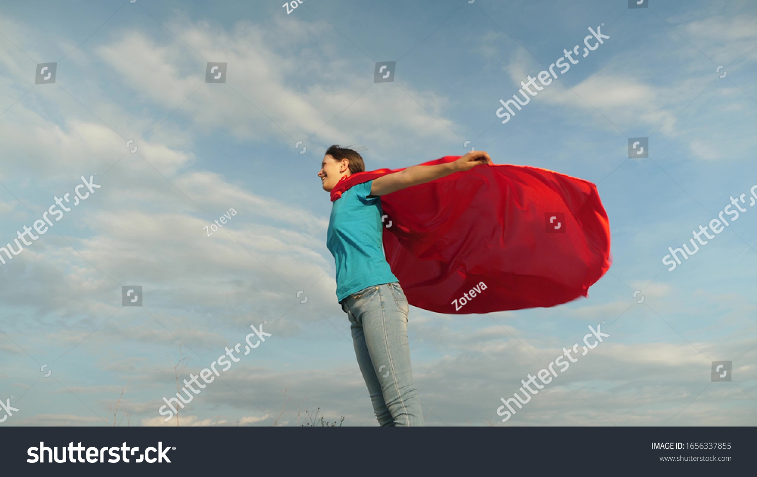 child plays in a red coat, dream of becoming a hero Superwoman. beautiful girl superhero standing on field in red cloak, cloak fluttering in the wind. Slow motion. girl dreams of becoming a superhero. #1656337855