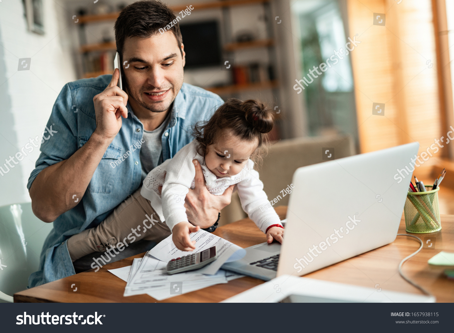 Young working father talking on the phone while babysitting his playful daughter at home. #1657938115