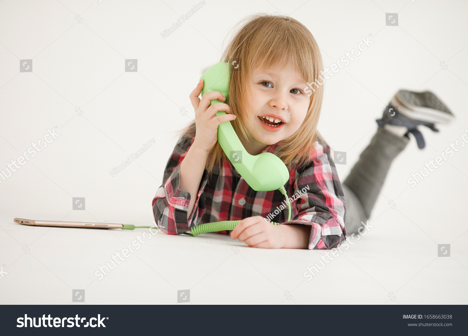 Emotional portrait of a cheerful and cheerful beautiful little girl looking with a smile out the window while talking on a telephone receiver connected to a smartphone, isolated on white background #1658663038