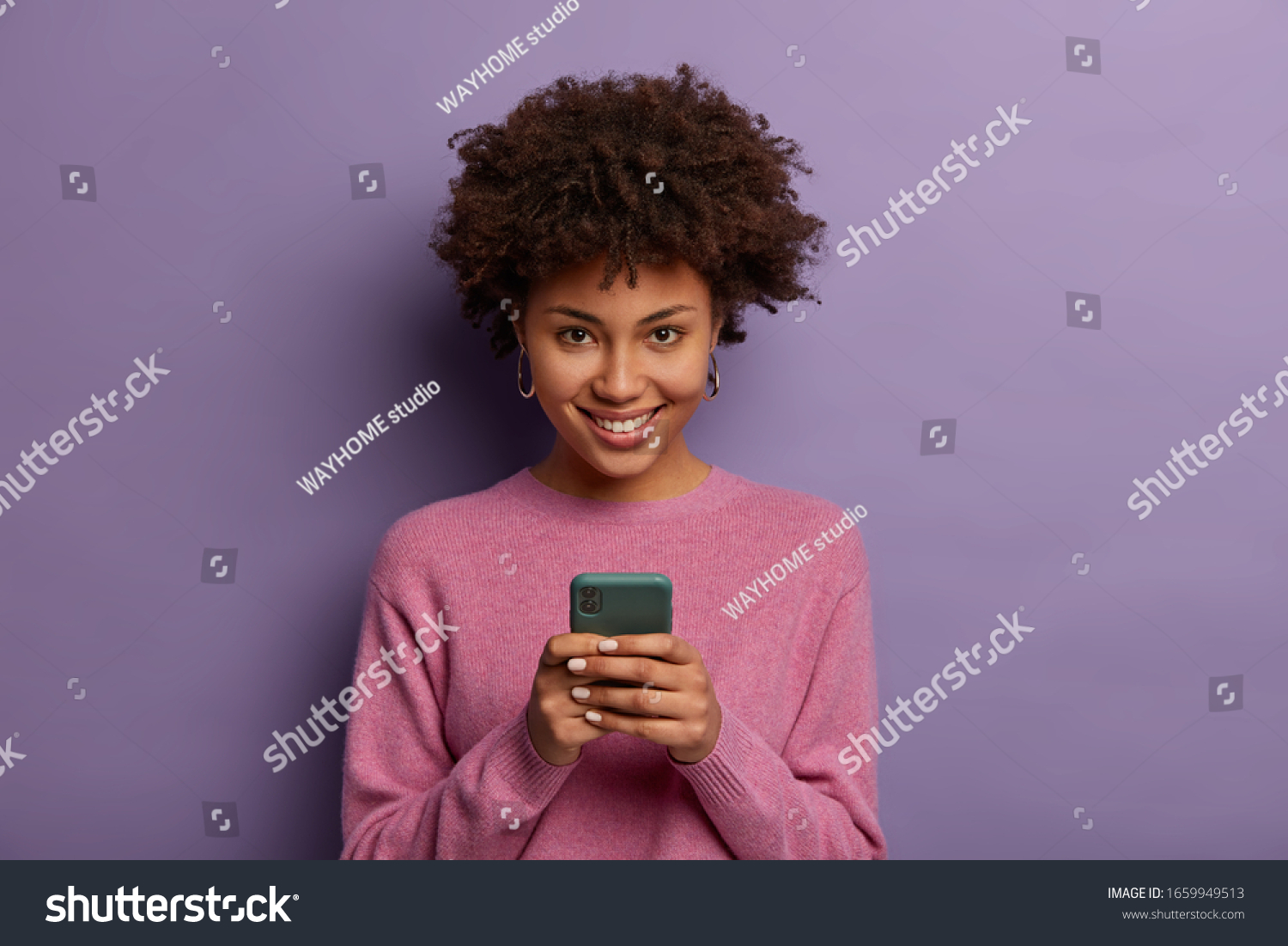 Portrait of lovely ethnic woman holds modern mobile phone, uses electronic device on surfing web, looks positively at camera, connected to wireless internet, wears casual sweater, poses indoor #1659949513
