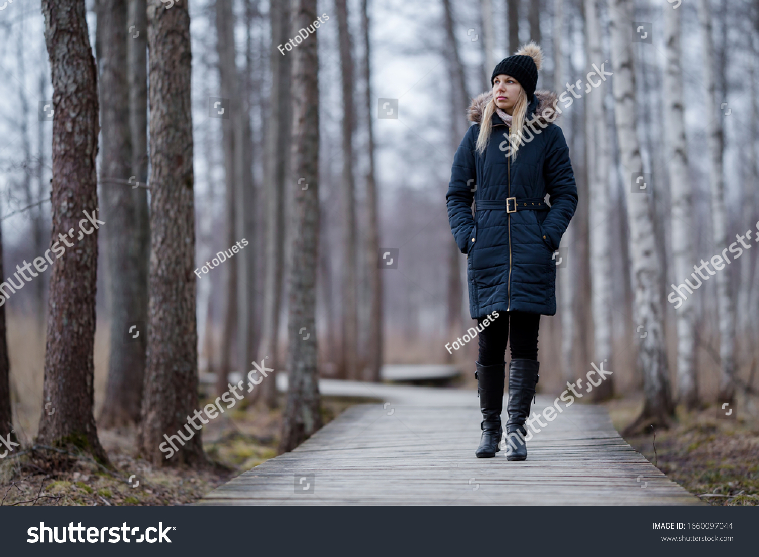 Young woman in dark warm clothes slowly walking on wooden trail at natural park. Cold overcast day. Spending time alone in nature. Peaceful atmosphere. Front view. 