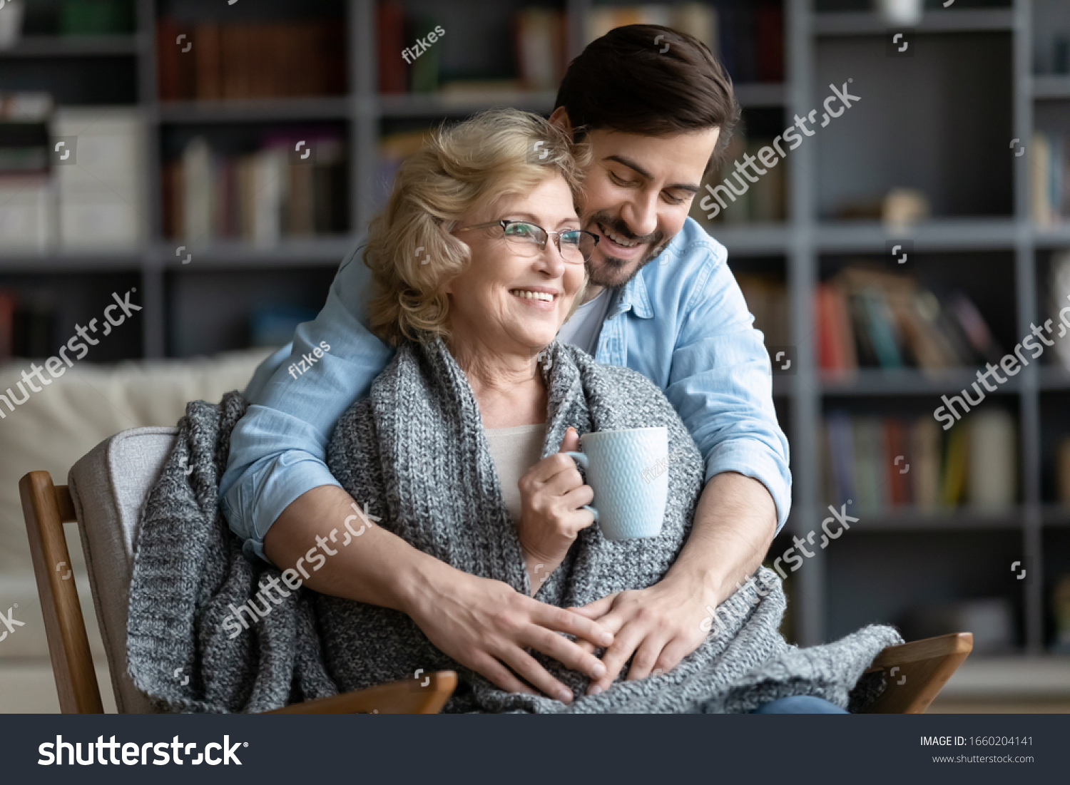 Loving young man take care of happy middle-aged mother sit in chair drinking tea enjoying family weekend in living room, thankful grown-up adult son hug show gratitude and affection to mature mom #1660204141