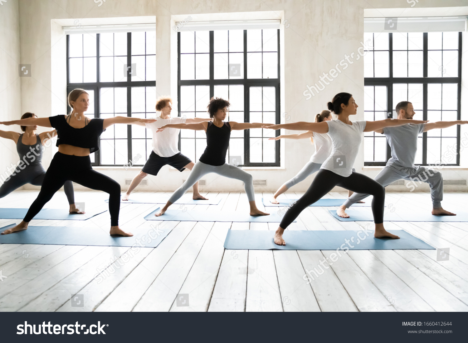 Group of multiracial people practicing yoga asanas, doing Warrior Two Virabhadrasana 2, work out indoors full length, posture increases stamina and flexibility, improves physical and mental endurance #1660412644