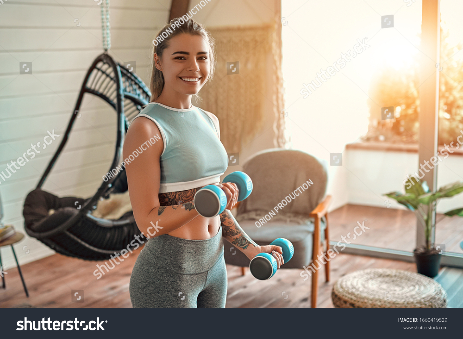 Determined woman losing weight at home and exercising with dumbbells. Sport and recreation concept. Beautiful woman in sportswear with blue dumbbells in her hands. #1660419529