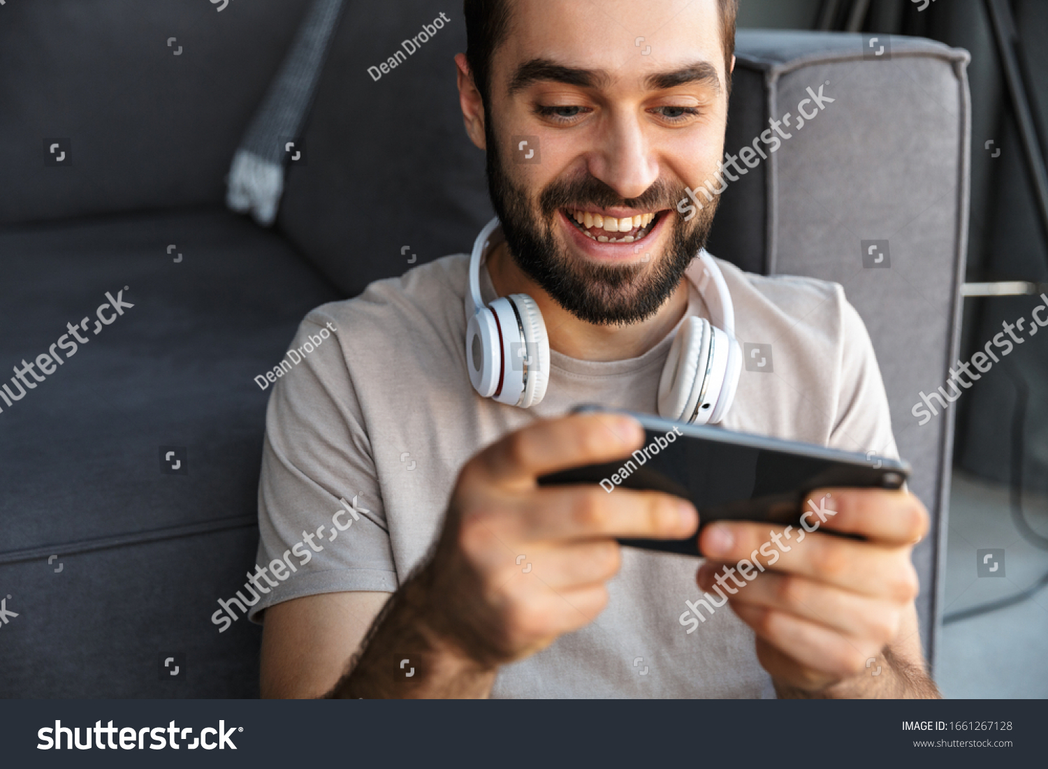 Photo of a happy optimistic young man indoors at home play games by mobile phone sit on floor. #1661267128