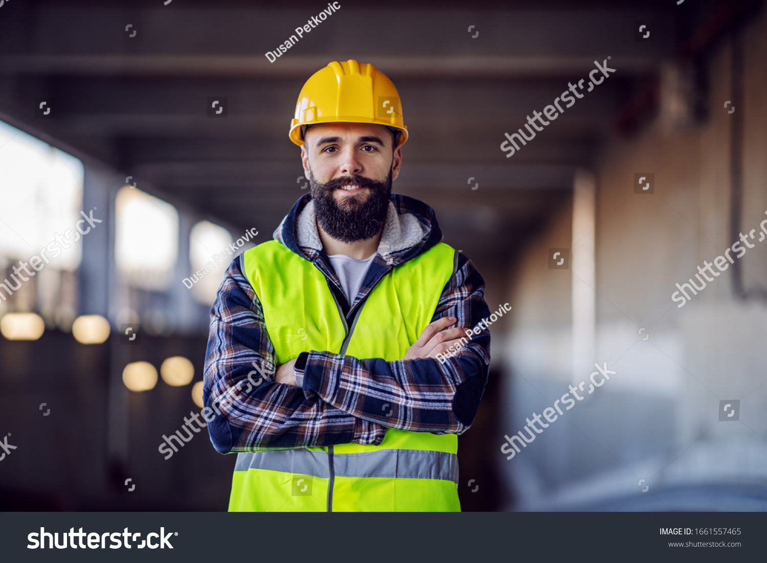 Cute Caucasian bearded construction worker with safety helmet on head in vest standing with arms crossed at construction site and looking at camera. #1661557465