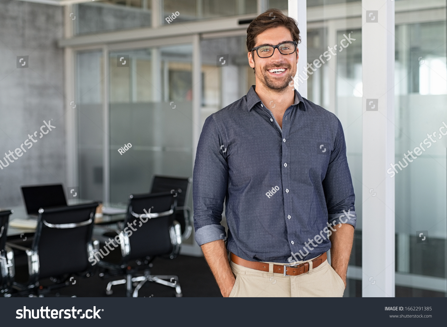 Portrait of young businessman wearing eyeglasses and standing outside conference room. Portrait of happy business man wearing spectacles and looking at camera with copy space. Satisfied proud man. #1662291385