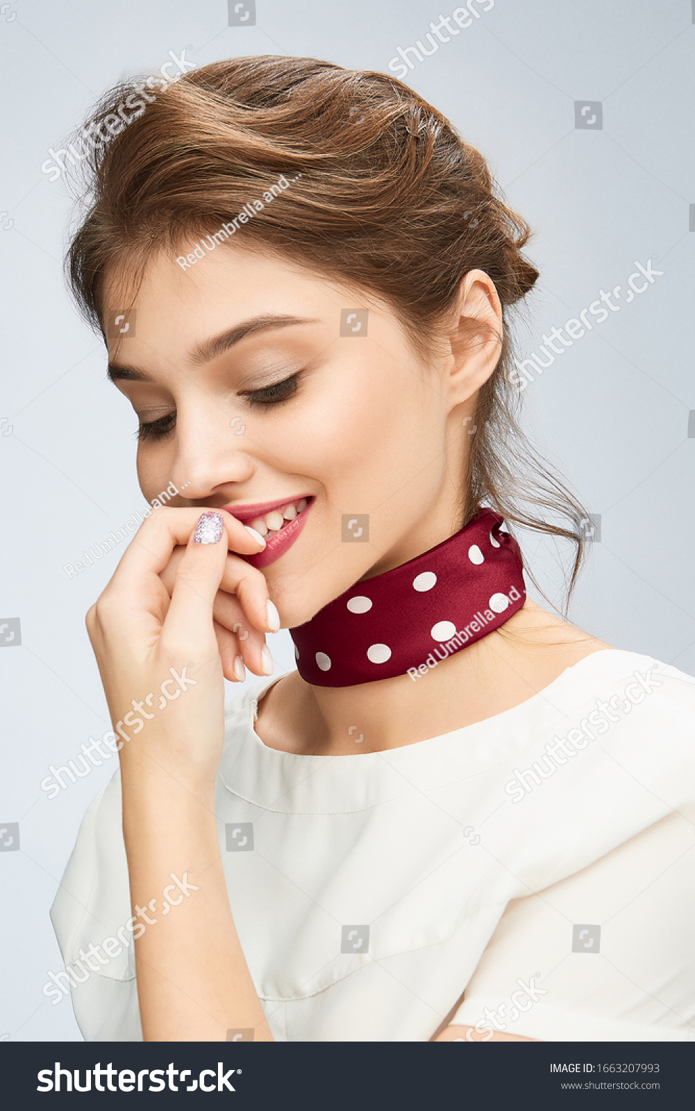 A chestnut-haired lady with careless hairdo in a white blouse is smiling and keeping her finger near the lips. A burgundy silk handkerchief with white polka-dot print is rounding the girl's neck. #1663207993