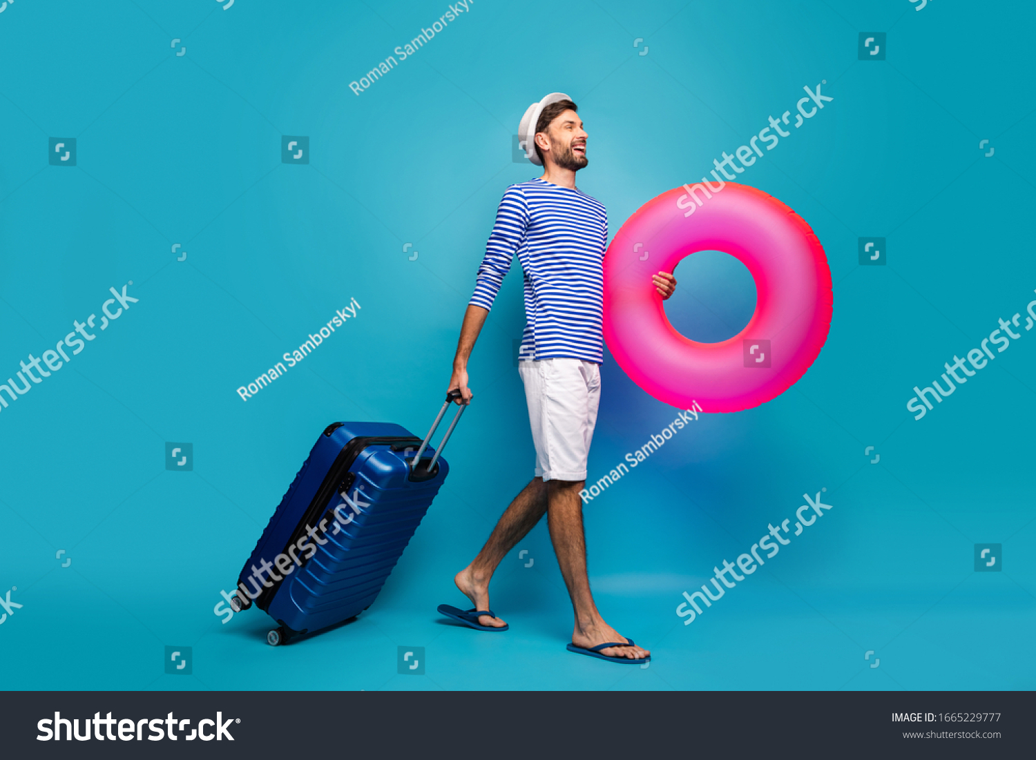 Full body profile photo of handsome guy traveler rolling case hold big pink lifebuoy want to see ocean wear striped sailor shirt cap shorts flip flops isolated blue color background #1665229777