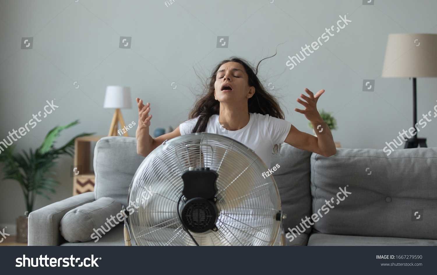Young Caucasian woman sit on couch in living room breathe fresh air from floor fan suffering from lack of conditioner, millennial girl use ventilator struggle with home heat, summer hot weather #1667279590