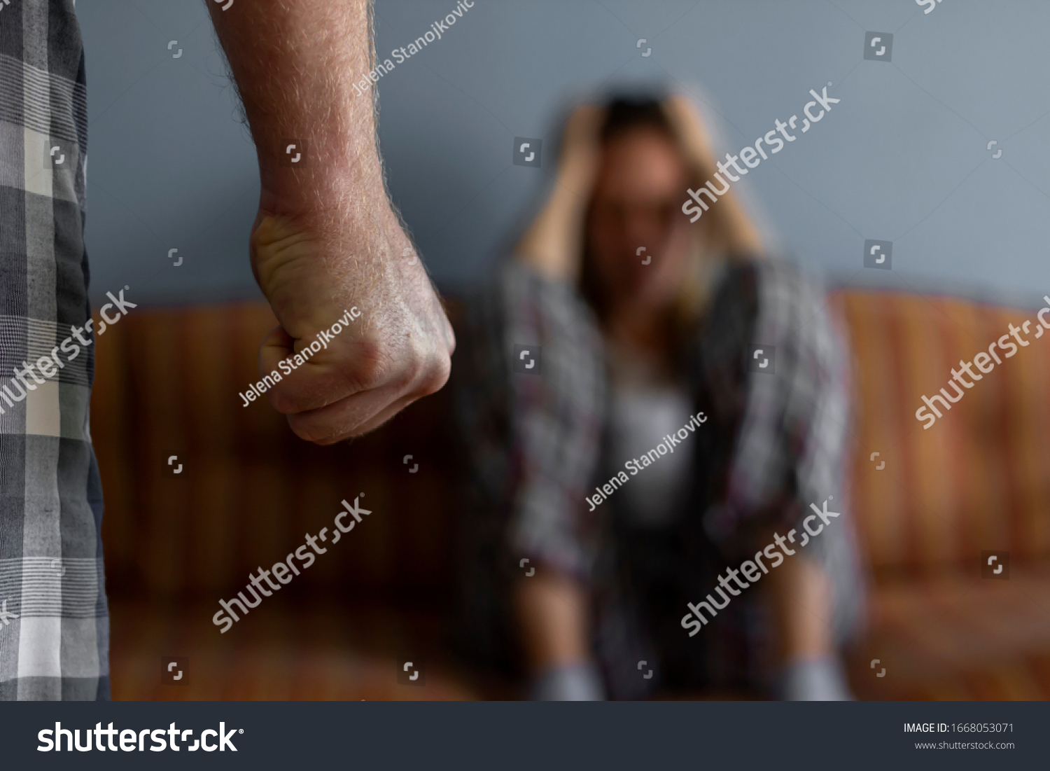 Photo of young woman sitting on sofa at home,focus is on man's fist in the foreground of the image.Home violence concept.Frightened woman and men's fist.Woman is victim of domestic violence and abuse. #1668053071