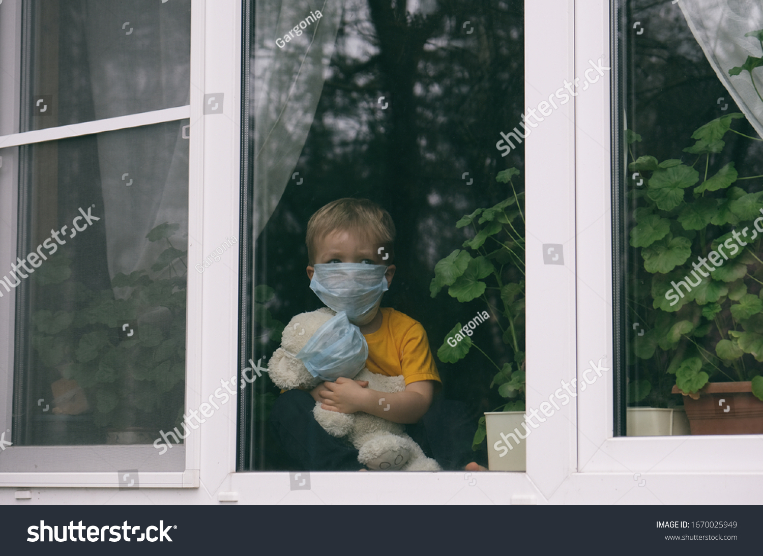Stay at home quarantine coronavirus pandemic prevention. Sad child and his teddy bear both in protective medical masks sits on windowsill and looks out window. View from street. Prevention epidemic. #1670025949