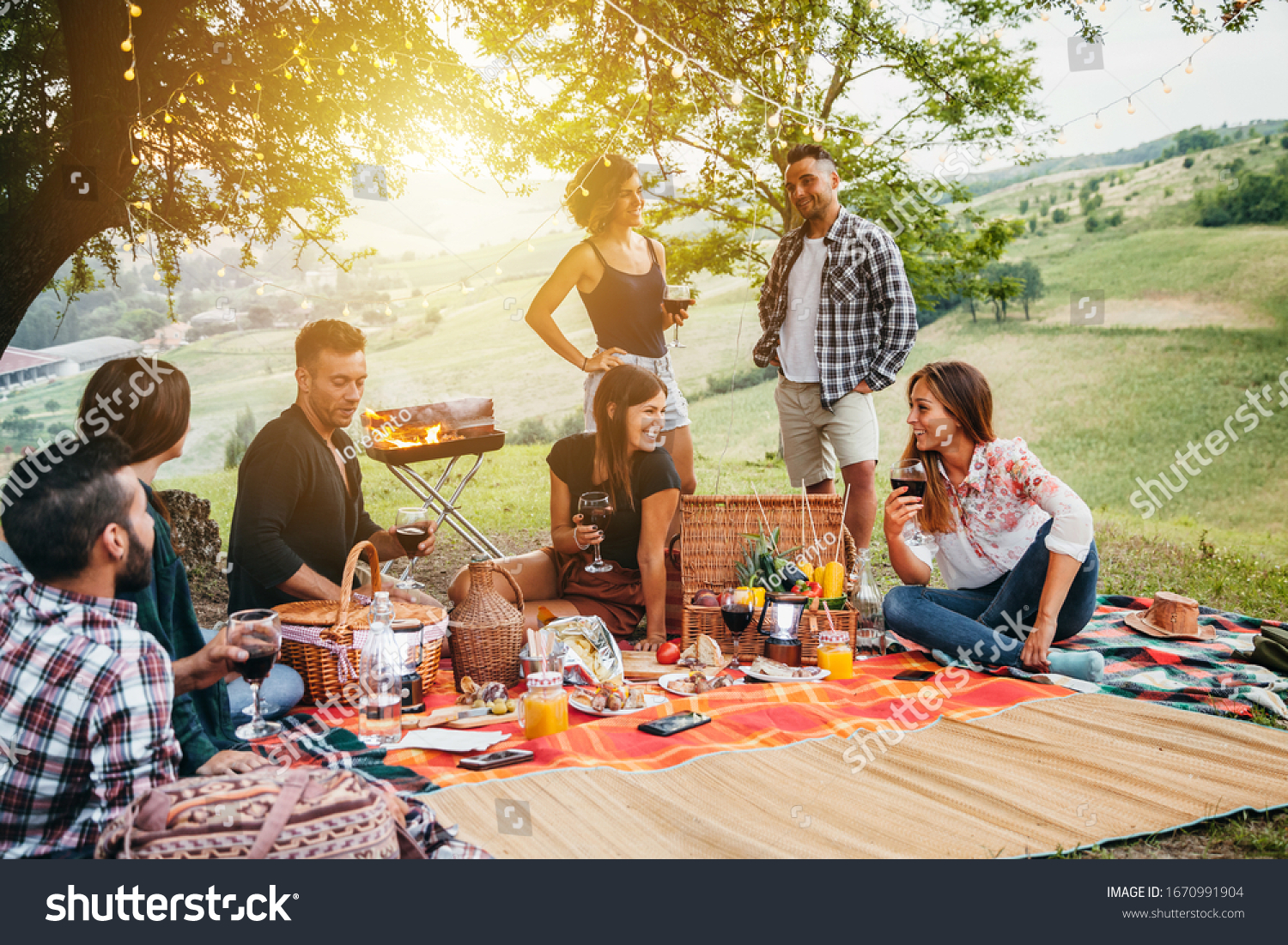 Picnic in the countryside. Group of young friends, at sunset on spring day are sitting on the ground in a park near trees. They drinking red wine and eating grilled meat with barbecue #1670991904