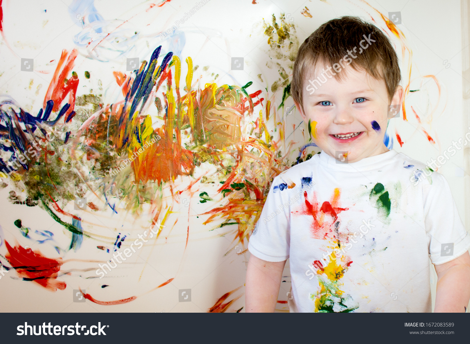 Cheerful happy blue-eyed child in a white T-shirt, painted and stained clothes. The boy looks into the frame, smiles. #1672083589