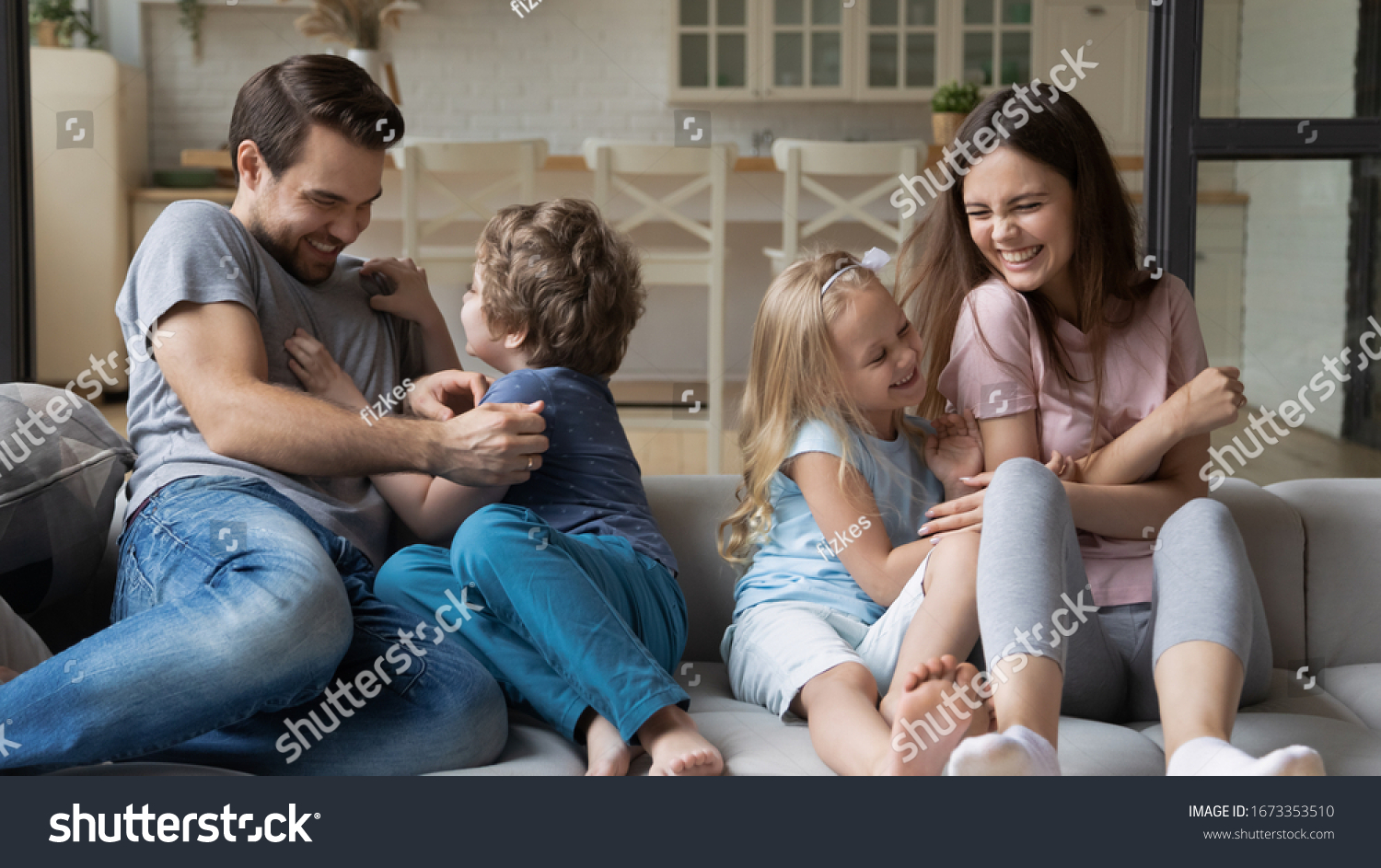 Playful young parents have fun play with happy little kids tickle laugh sitting on couch in living room, overjoyed family enjoy funny childish activity or game with small children on weekend at home #1673353510