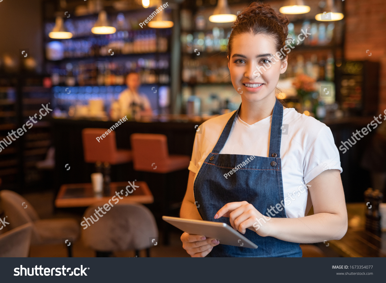 Young smiling waitress in apron and t-shirt standing in front of camera while using touchpad and meeting guests in cafe #1673354077