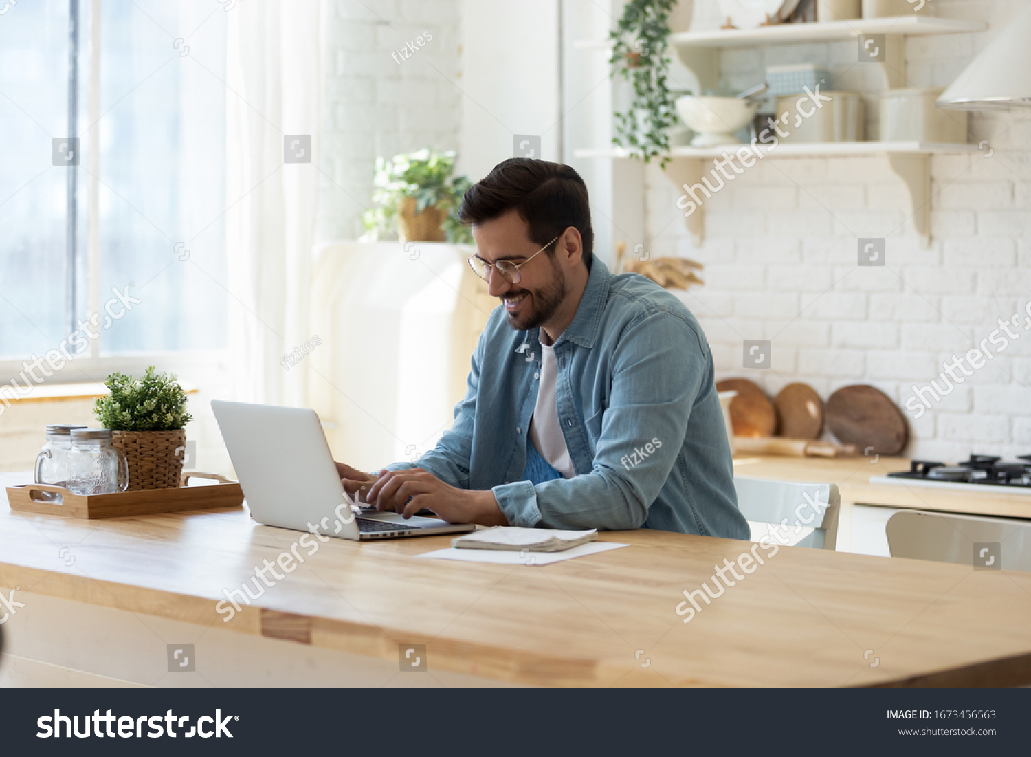Smiling young man working on laptop in modern kitchen, checking email in morning, writing message in social network, happy young male using internet banking service, searching information #1673456563