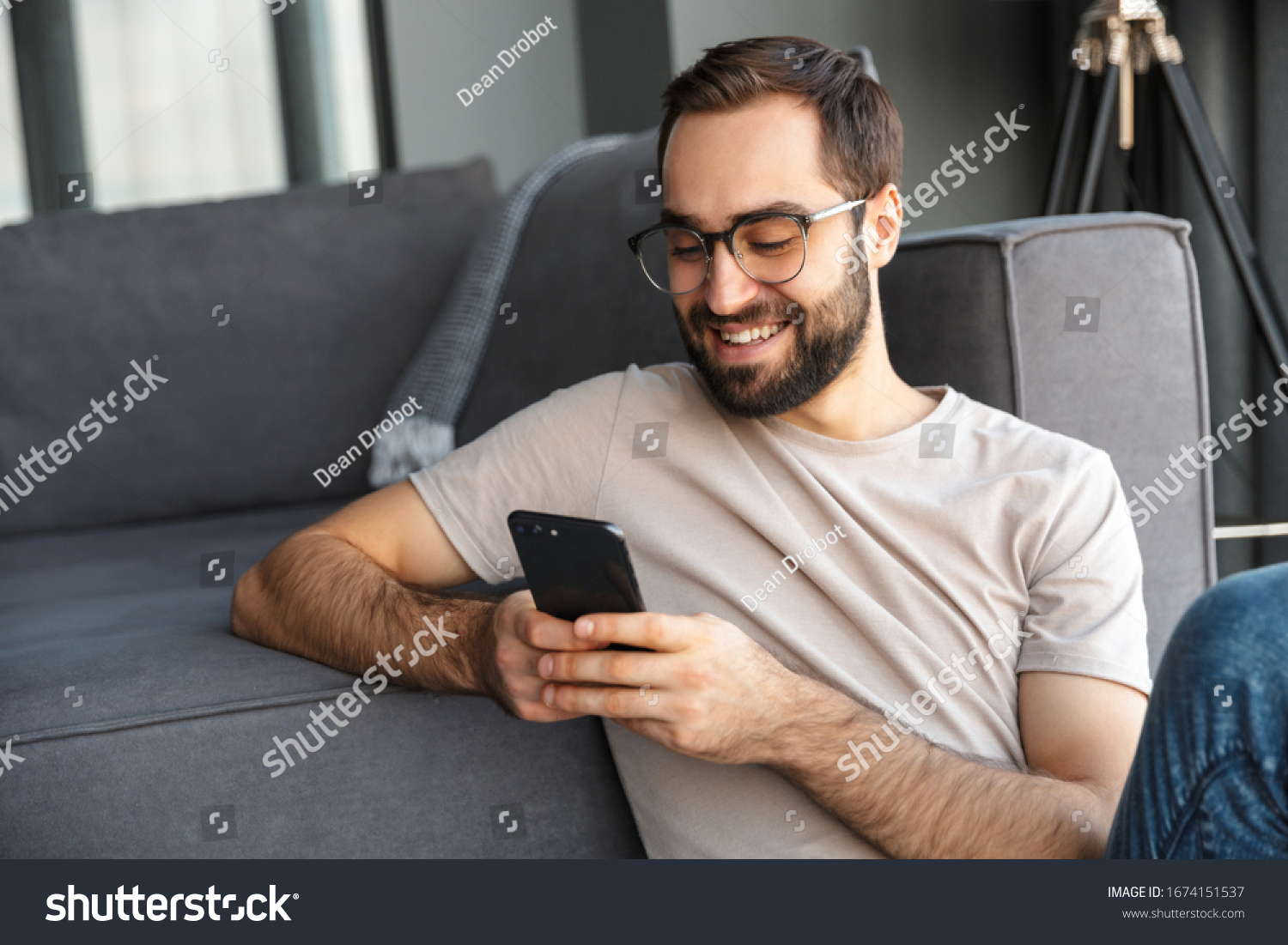 Attractive smart young man sitting on a floor in the living room, using mobile phone #1674151537