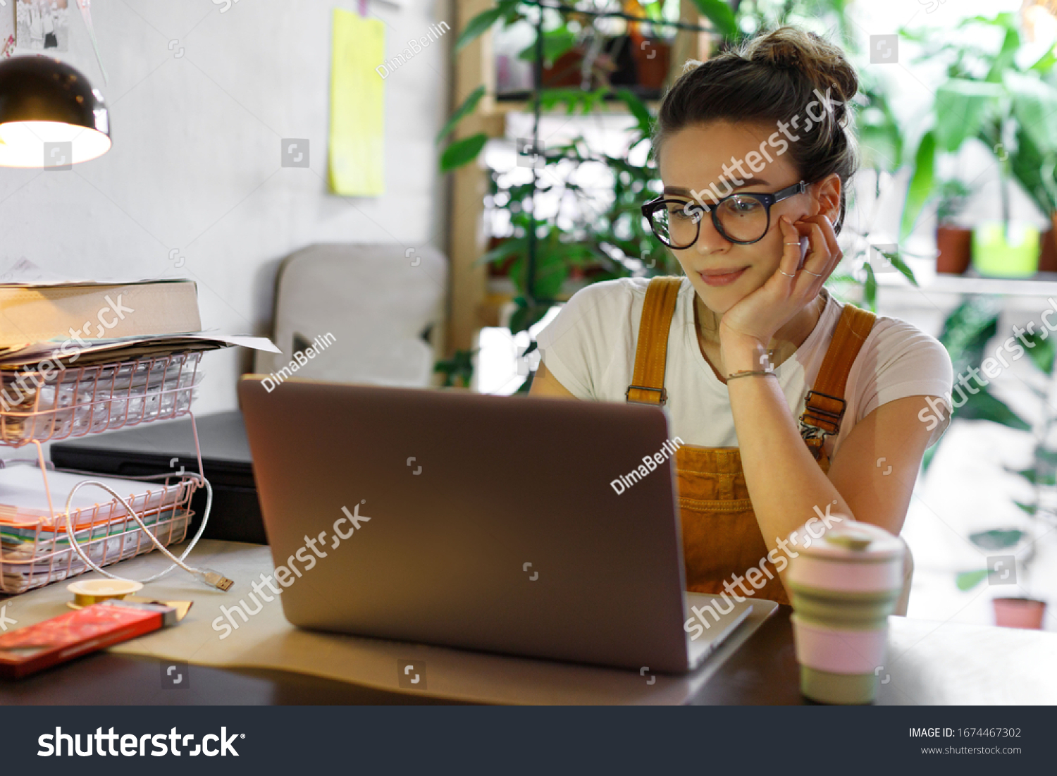 Young female gardener in glasses using laptop, communicates on internet with customer in home garden/greenhouse, reusable coffee/tea mug on table.Cozy office workplace, remote work, E learning concept #1674467302