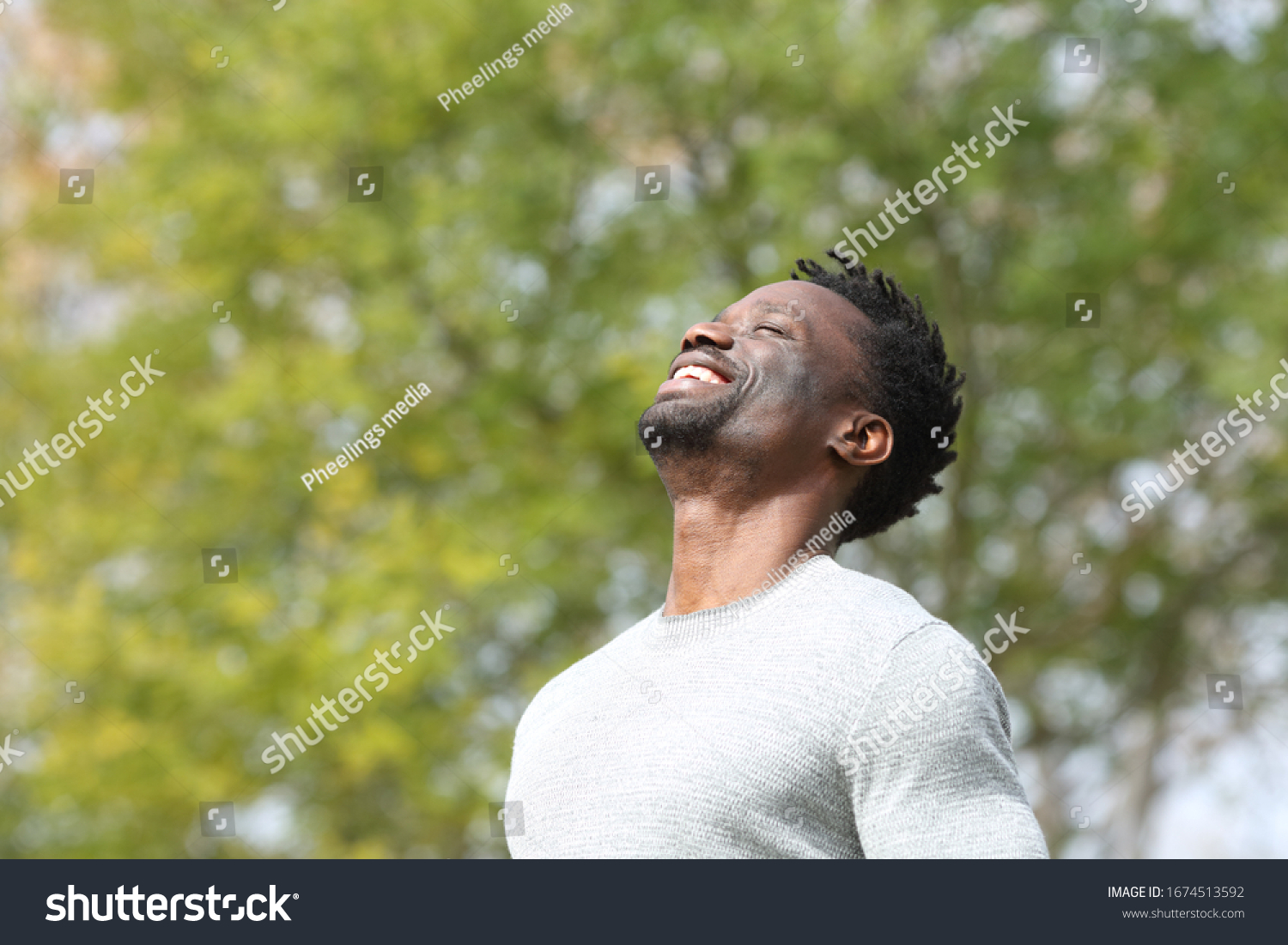 Happy black man breathing deeply fresh air in a park with a green tree in the background a sunny day #1674513592