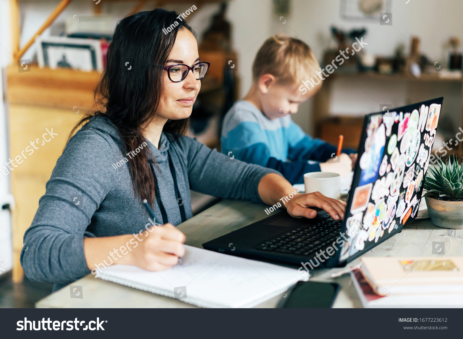 Concept of work from home and home family education. Mom and son are sitting at the desk. Business woman works on the Internet in a laptop, a child writes in a notebook. #1677223612