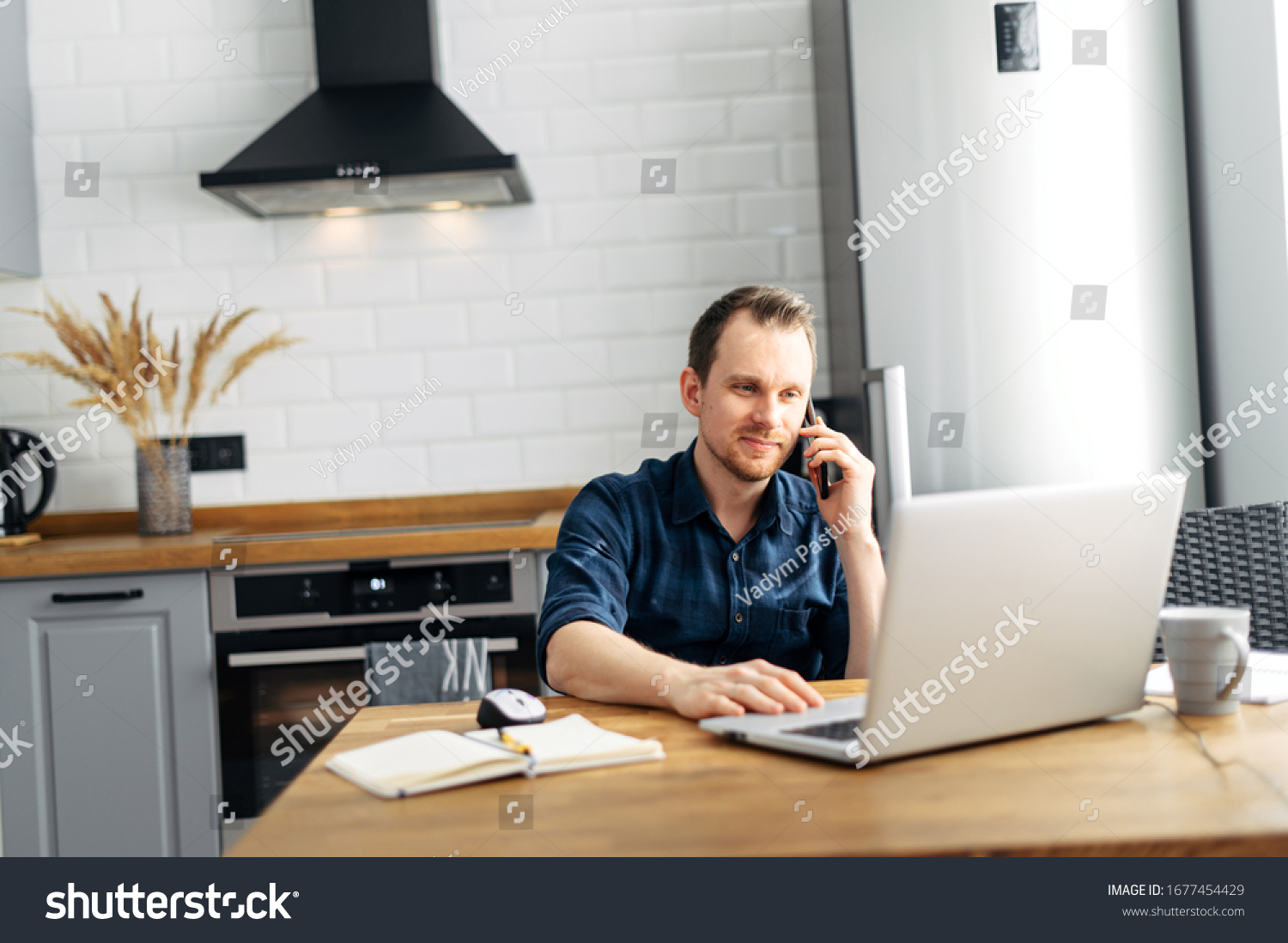 Freelancer in the kitchen at home. A young attractive guy uses a laptop for work, he sends emails and answers calls #1677454429