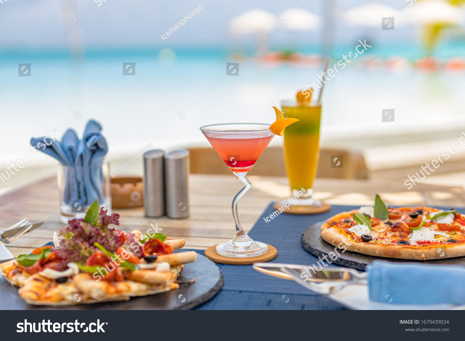 Pizza and tropical cocktail with beautiful sunny vacation infinity swimming pool ocean view. Beach restaurant, sea view, glasses, plates, food, tropical cocktail, blur loungers, chairs, sun umbrellas #1679439034