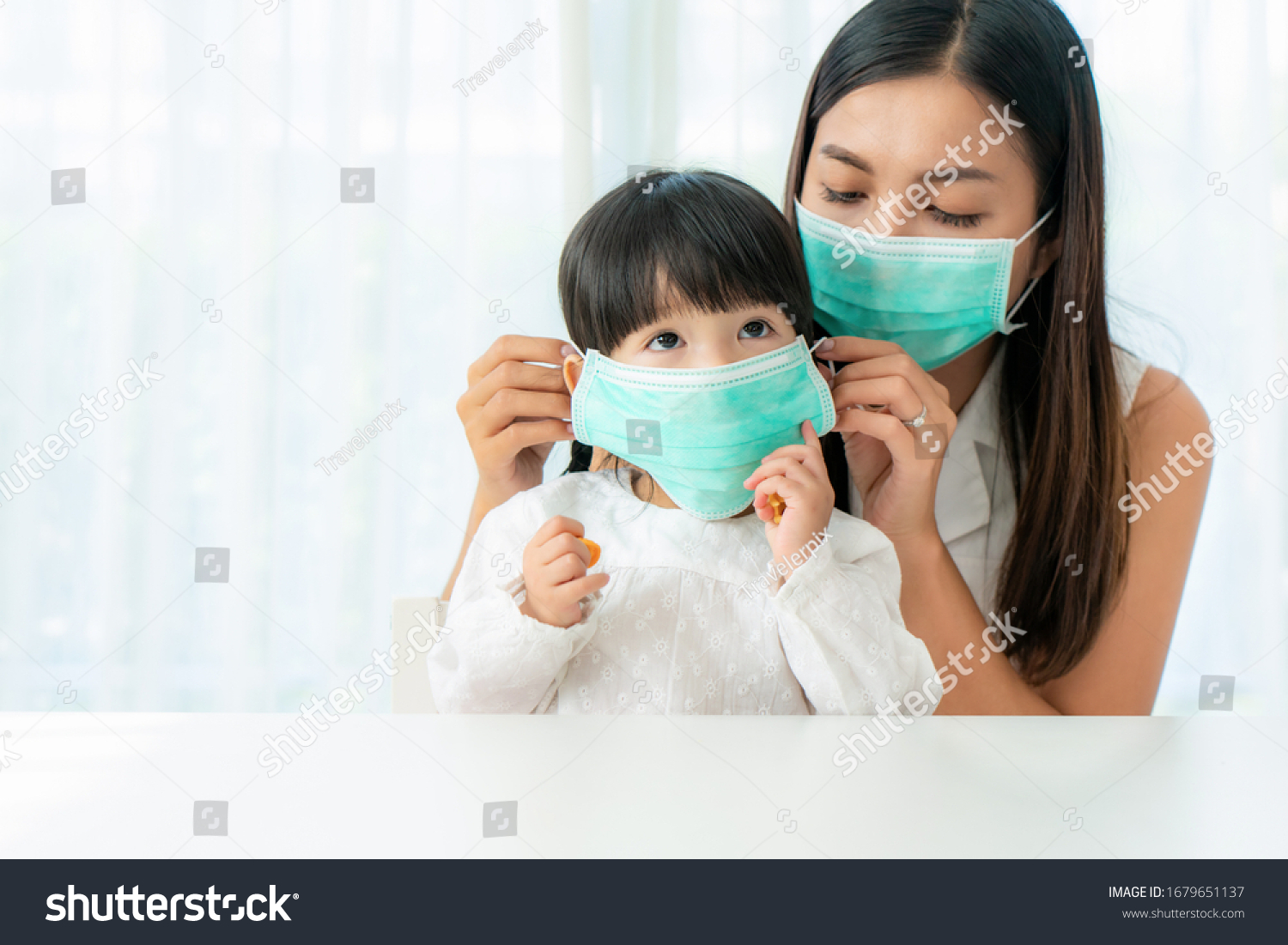 Asian mother wearing wearing to her daughter healthy face mask sitting in living room at home to prevent PM2.5 dust, smog, air pollution and COVID-19. Healthcare concept. #1679651137