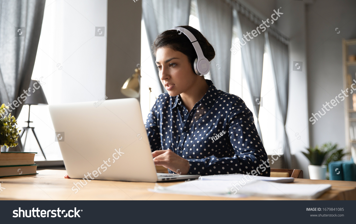 Busy young Indian woman wearing headphones working on laptop, looking at screen, making video call, engaged in conference, focused student listening to lecture, learning language online #1679841085