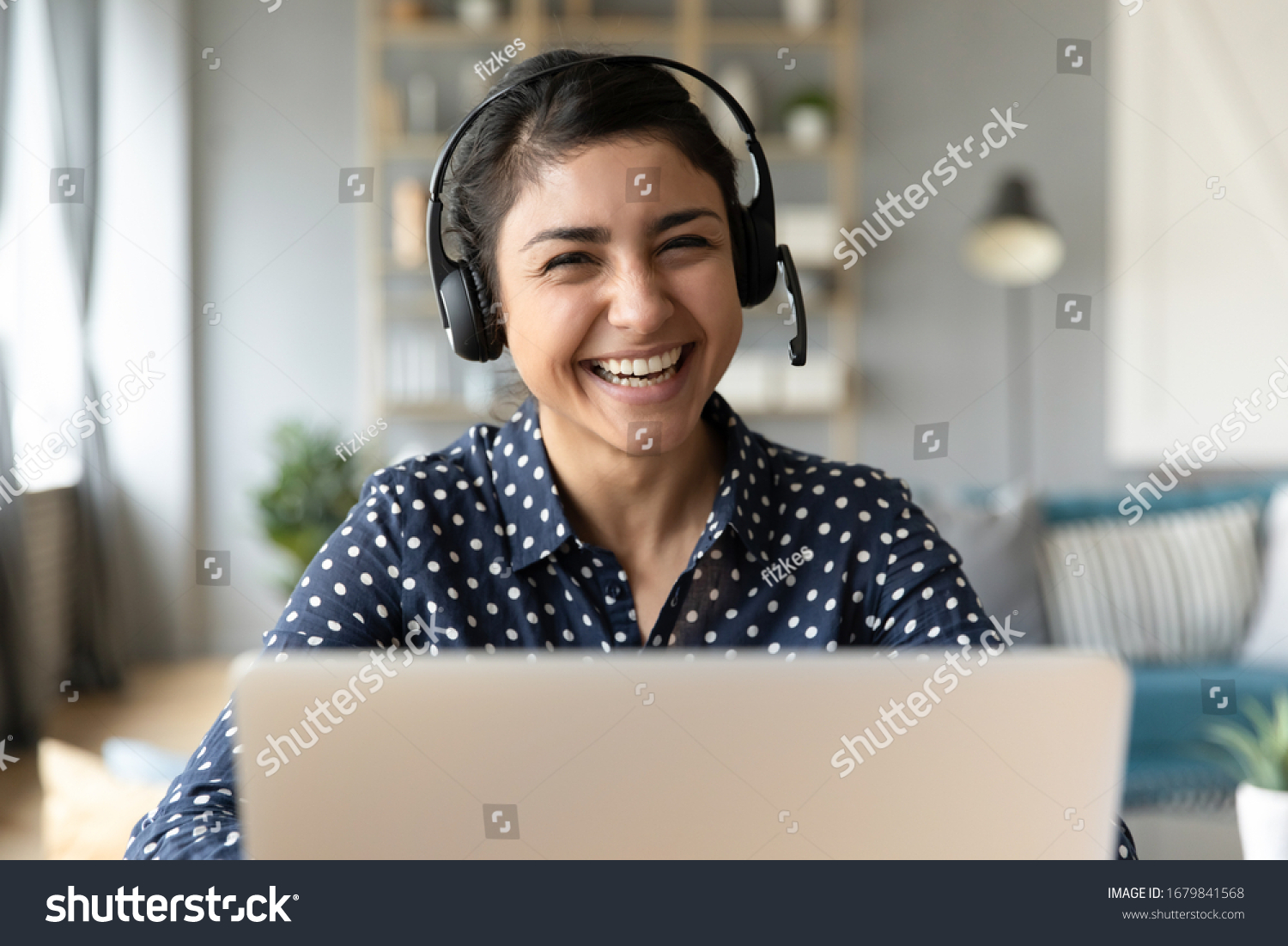 Head shot portrait smiling Indian woman wearing headphones posing for photo at workplace, happy excited female wearing headset looking at camera, sitting at desk with laptop, making video call #1679841568
