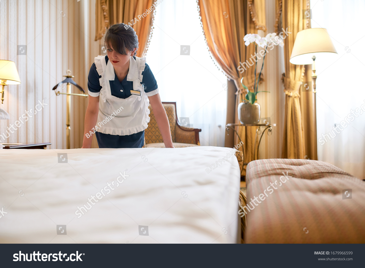 Hotel maid in uniform changing linen and making bed in luxury hotel room. Room service concept. Horizontal shot #1679966599