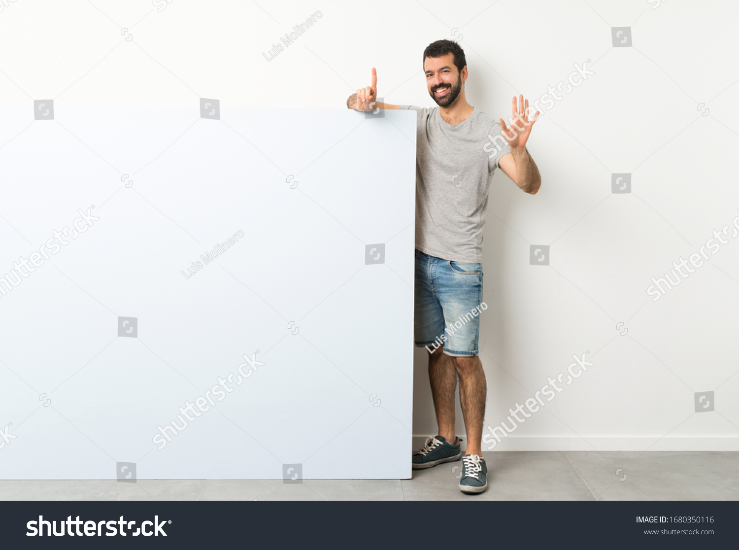 Young handsome man with beard holding a big blue empty placard counting six with fingers #1680350116