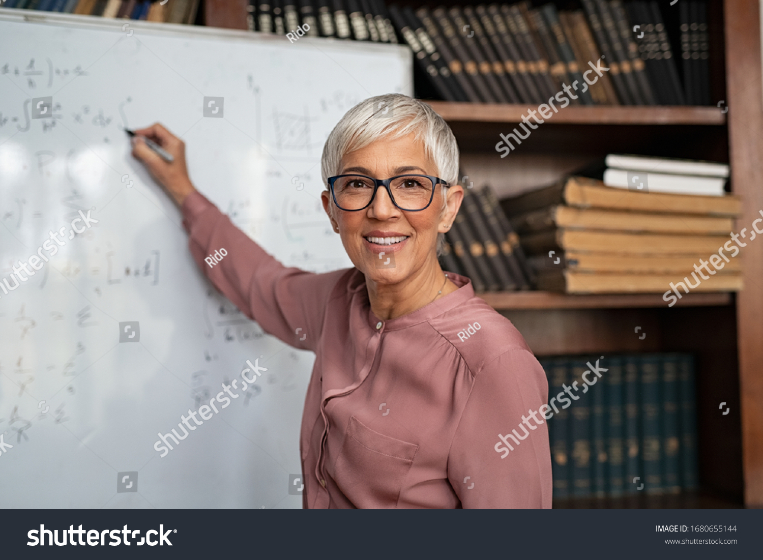 Portrait of happy mature professor teaching mathematics to students in a library. Senior smiling woman solving math problem while writing on white board. Portrait of tutor looking at camera. #1680655144