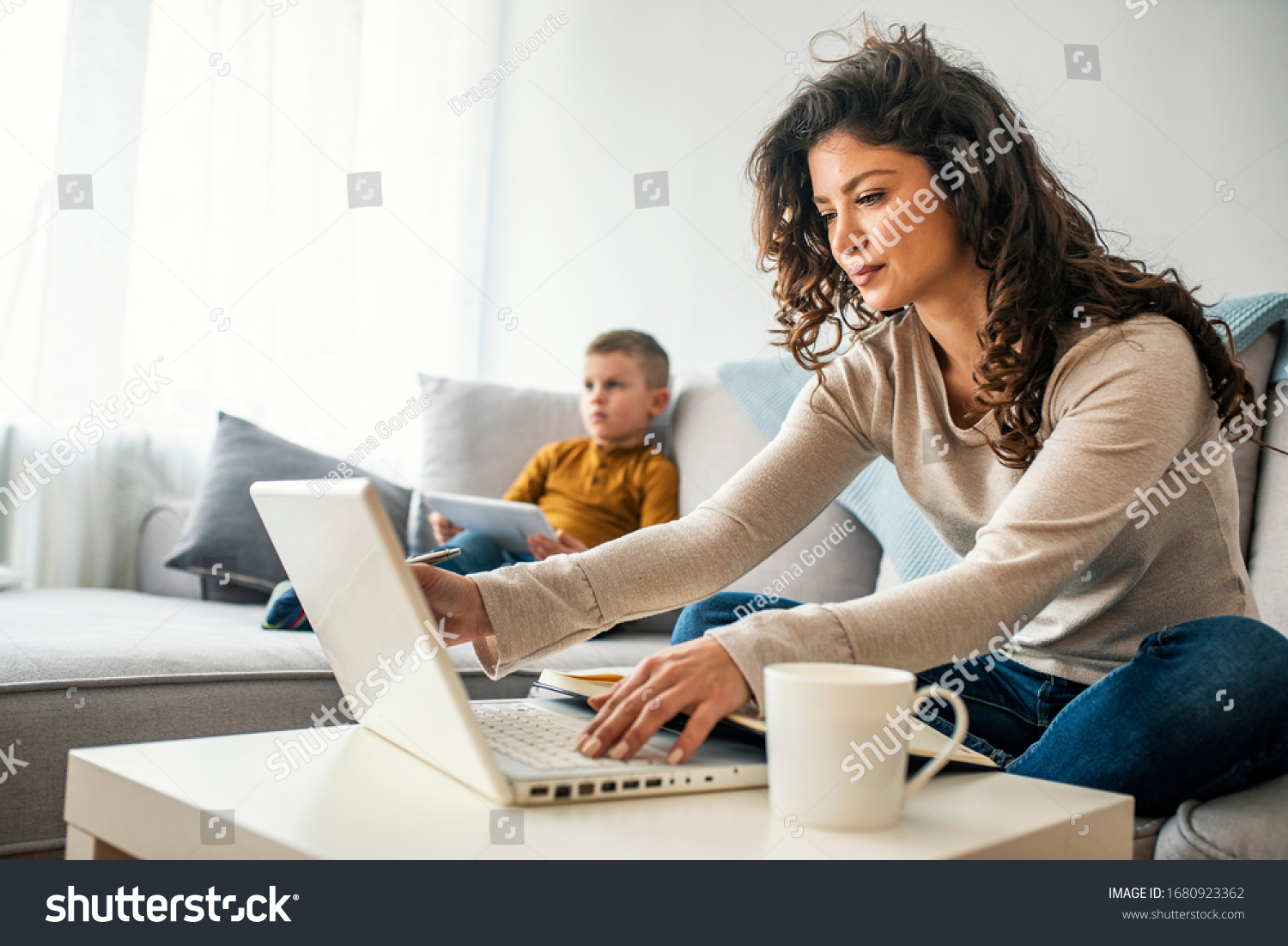 Smiling mom working at home with her child on the sofa while writing an email. Young woman working from home, while in quarantine isolation during the Covid-19 health crisis #1680923362