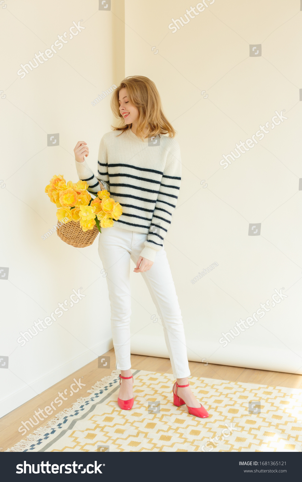 Shoot fashion campaign story. Charming model staying, wearing white textile sweater with stripes and creamy pants. Girl holding bunch of tulips flowers. Colorful carpet on floor. White Background.  #1681365121