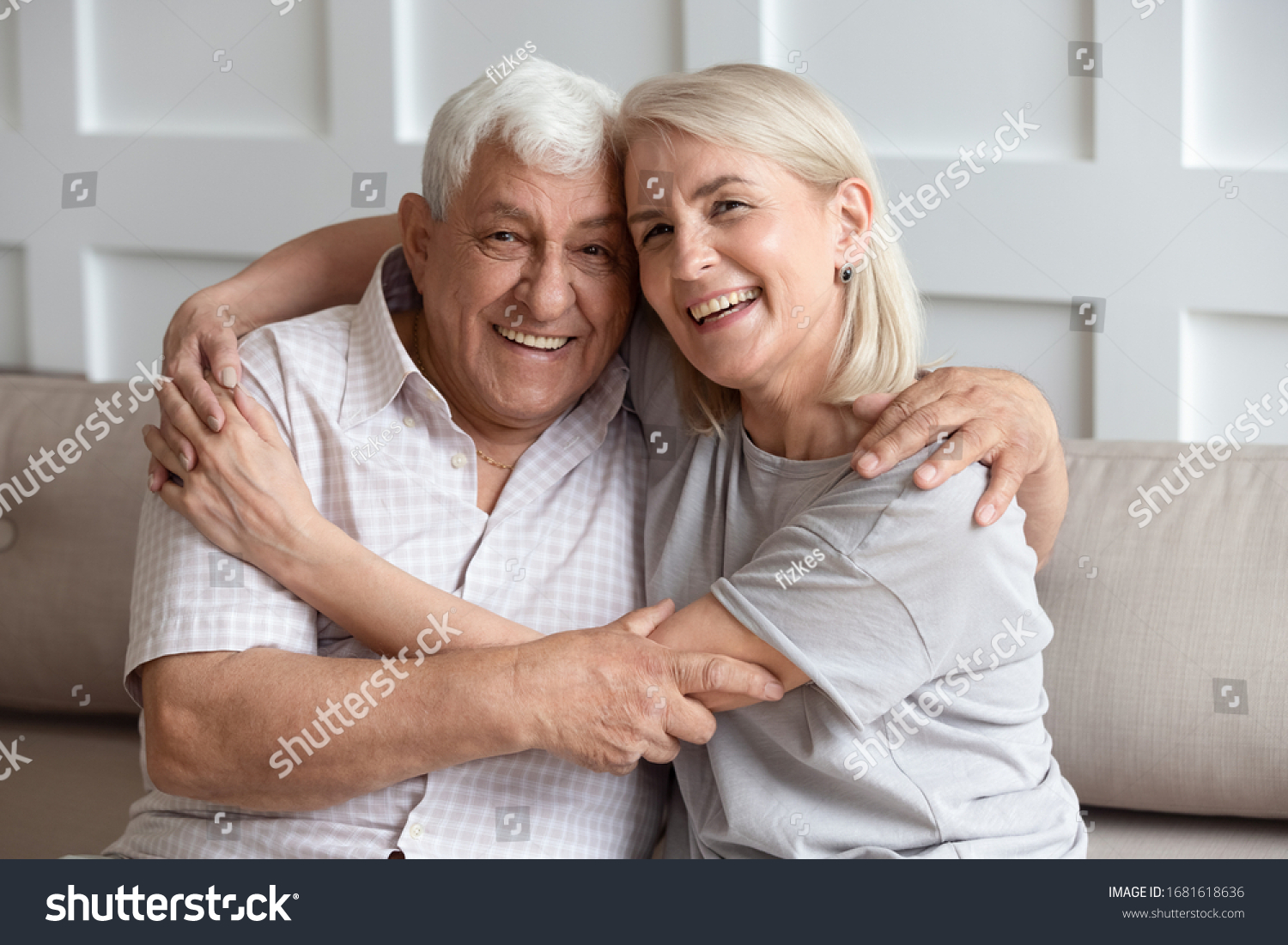 Head shot portrait older wife and husband with healthy toothy smiles hugging, looking at camera, sitting on cozy sofa at home, happy adult middle-aged daughter embracing mature father, family photo #1681618636