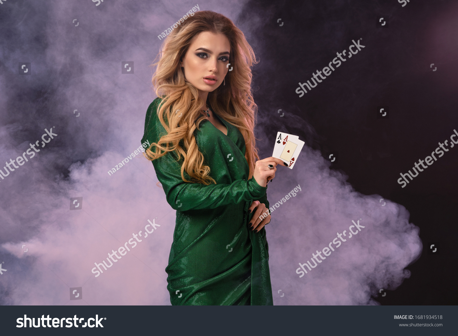 Blonde model with make-up, in green dress and jewelry. Put hand on hip, showing two aces, posing on black smoky background. Poker, casino. Close-up #1681934518