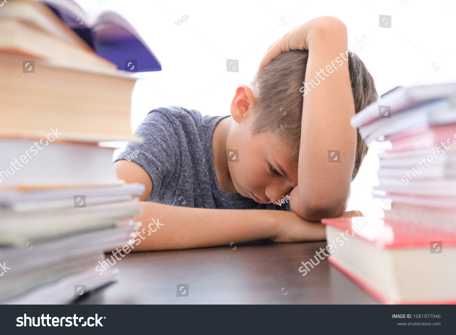 Tired disappointed schoolboy lowering his head sitting among pile of books, textbooks, school exercise books on his desk at home #1681977946
