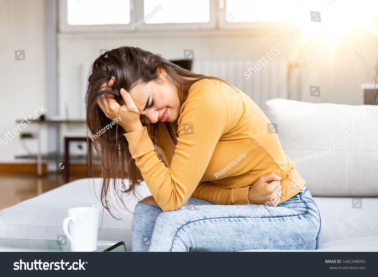 Young sick woman with hands holding pressing her crotch lower abdomen. Medical or gynecological problems, healthcare concept. Young woman suffering from abdominal pain while sitting on sofa at home #1682348995