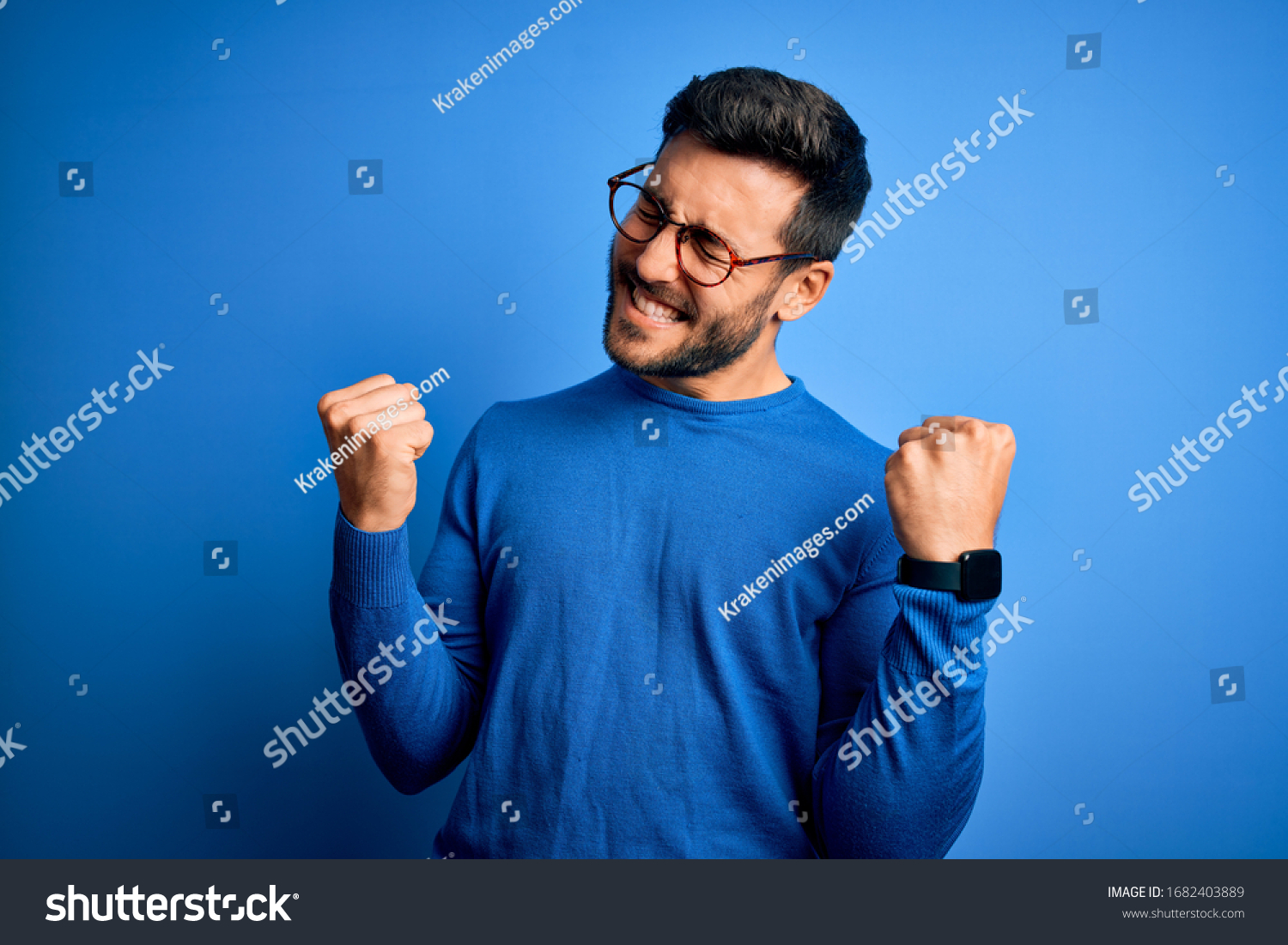 Young handsome man with beard wearing casual sweater and glasses over blue background very happy and excited doing winner gesture with arms raised, smiling and screaming for success. Celebration #1682403889