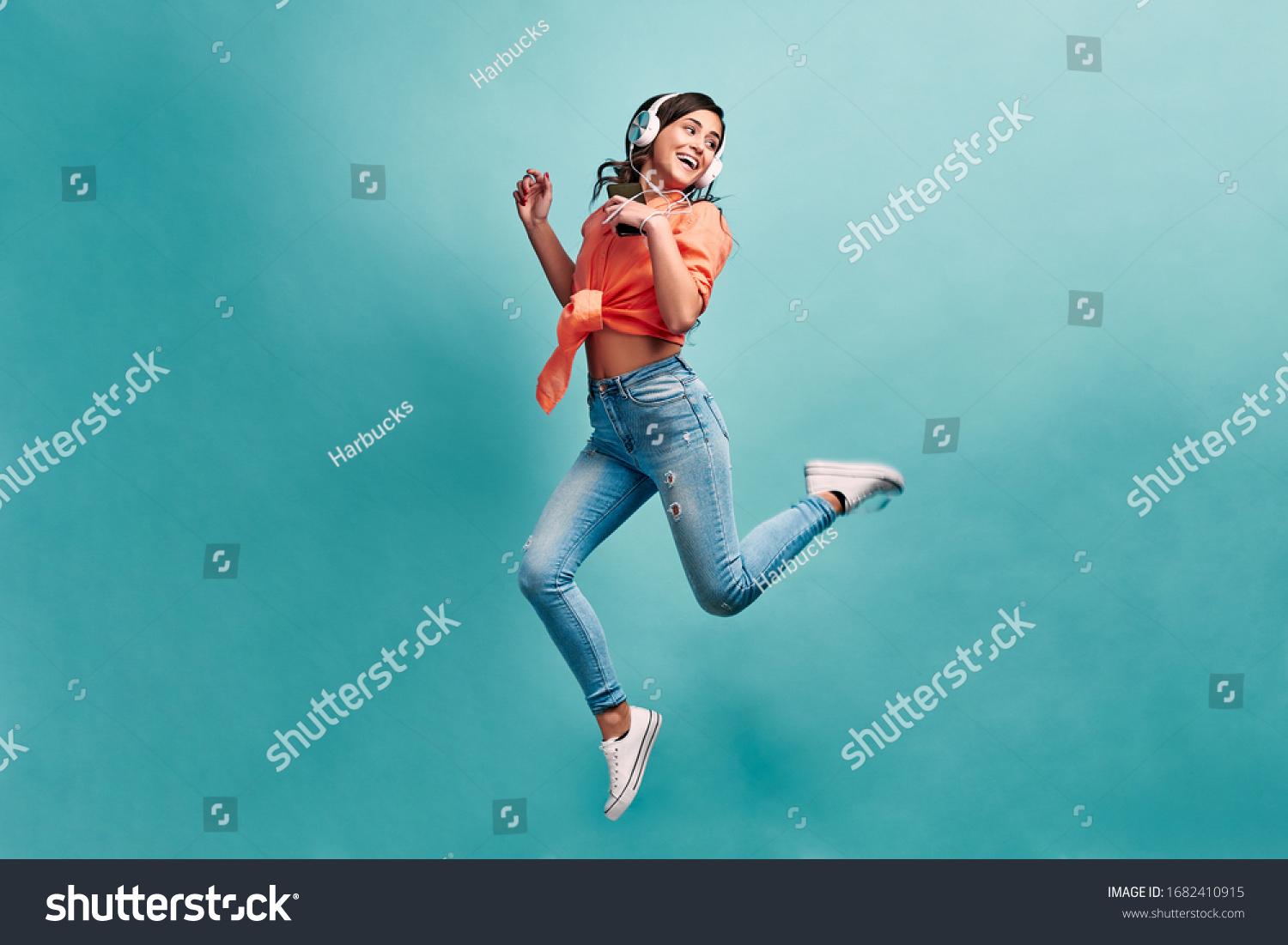 Young beautiful energy girl with white headphones listening to music laughs and jump on blue background in studio and looks away.Dressed in an orange shirt and light jeans, holding a phone. #1682410915