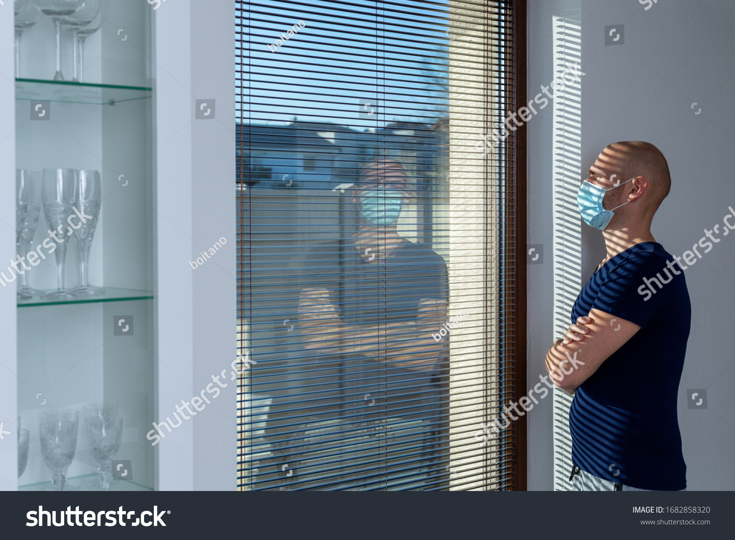 Home quarantine (self-isolation) because of the Coronavirus disease (COVID-19). Infected man with the ear-loop mask on his face looks out of the window. Man with protective face mask. Infection theme. #1682858320